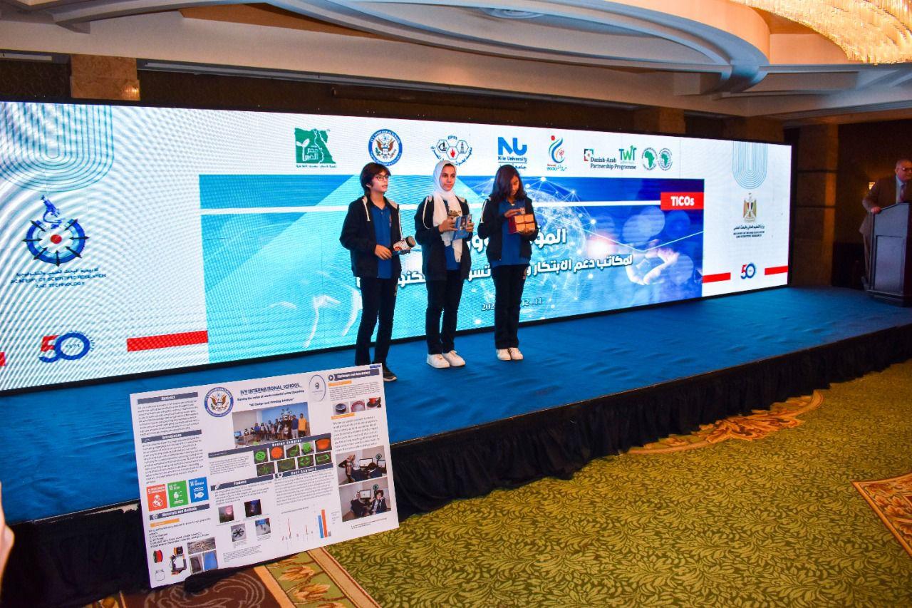 Three individuals on a stage presenting their project at the IVY STEM International School Academic Conference. The poster contains text and images elaborating on a particular project or research. The event is sponsored or organized by multiple entities, as indicated by the logos and emblems displayed on the large screen. The presence of Arabic text suggests the involvement of Arabic-speaking participants or the event taking place in an Arabic-speaking country.